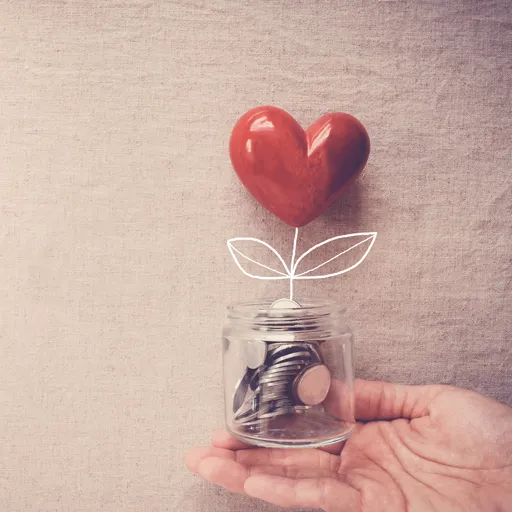 Eine Hand hält ein Glas mit Münzen, aus dem eine gezeichnete Pflanze mit einem roten Herzen als Blüte wächst – Symbolbild für Spenden.