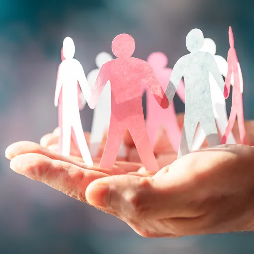 Eine offene Hand hält eine Gruppe von Papierfiguren in weiß, rosa und pink, die sich an den Händen halten, vor unscharfem Hintergrund – Symbolbild für Zusammenhalt und Unterstützung.